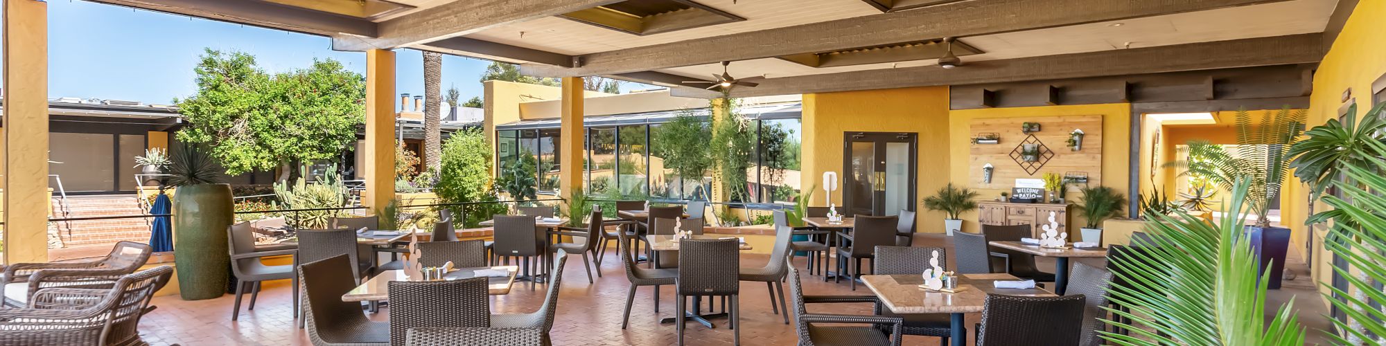 A spacious, outdoor dining area with wicker tables and chairs, plants, and yellow walls. The area is covered, offering a relaxed atmosphere.