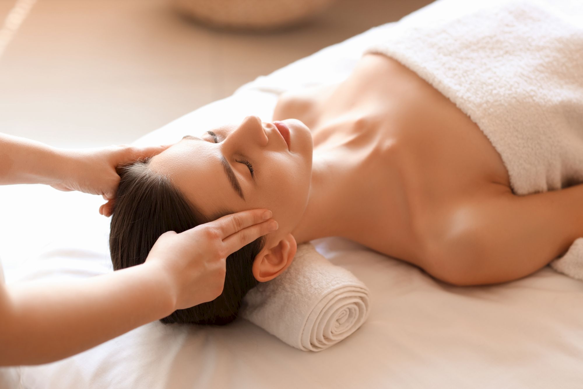 A person is receiving a head massage while lying on a spa bed with a towel draped over them and a rolled towel under their head.