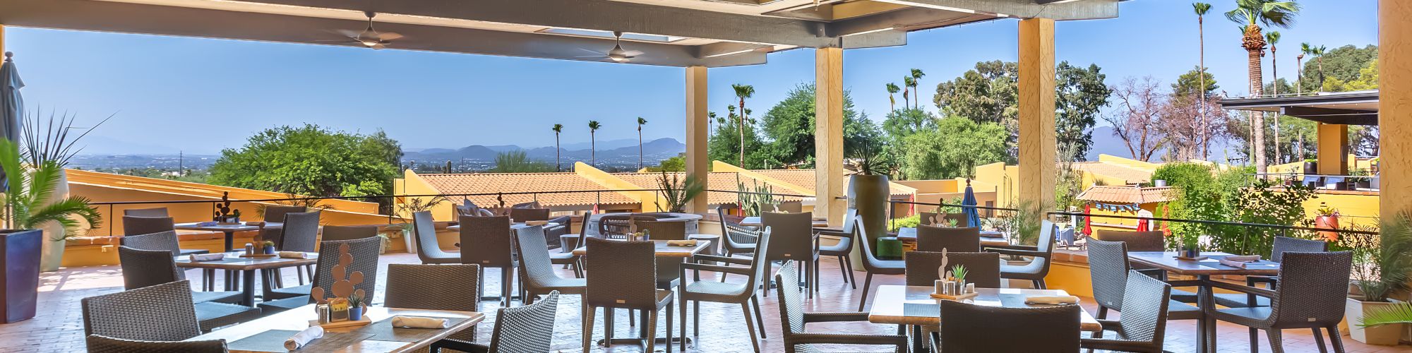 The image depicts an outdoor restaurant seating area with empty tables and chairs under a covered patio, overlooking a scenic view.