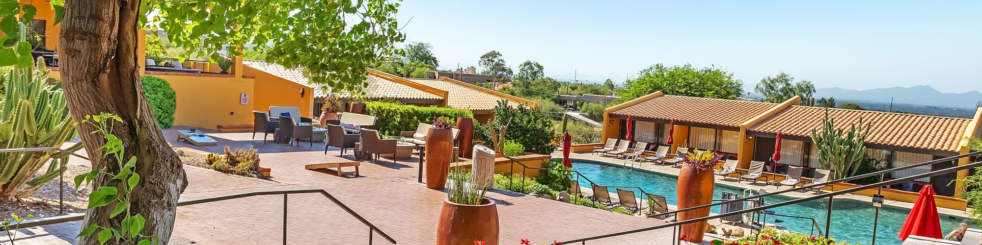 An outdoor pool area with sun loungers, surrounded by colorful flowers and greenery, and buildings with tiled roofs in the background.