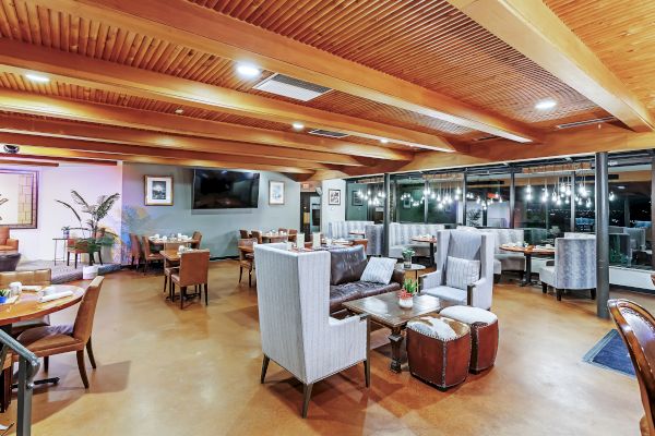 A modern, well-lit restaurant interior with wooden ceilings and a mix of table and lounge seating, decorated with plants and wall art.