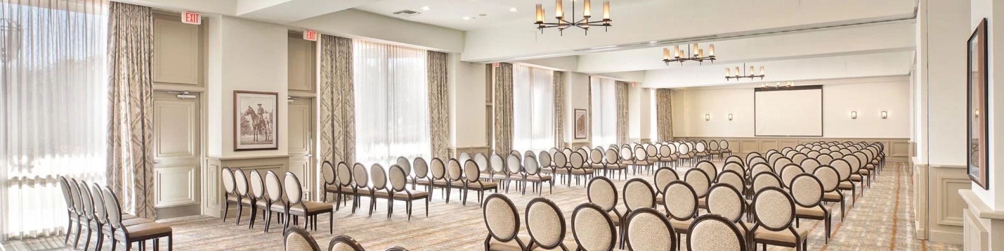 This image shows an empty conference room with rows of chairs facing a screen, chandeliers, and large windows with drapes in the background.