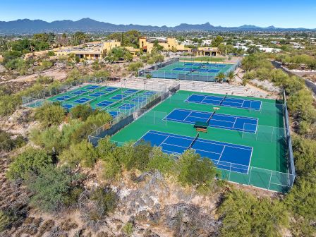 The image shows a complex of tennis and pickleball courts surrounded by desert terrain, with a backdrop of mountains and buildings in the distance.