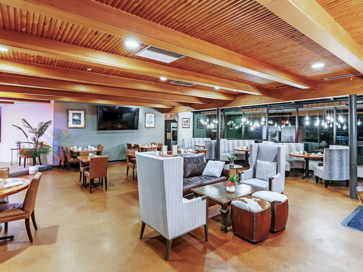 The image shows a spacious, modern restaurant with wooden ceilings, various seating arrangements, and large windows revealing a nighttime view.