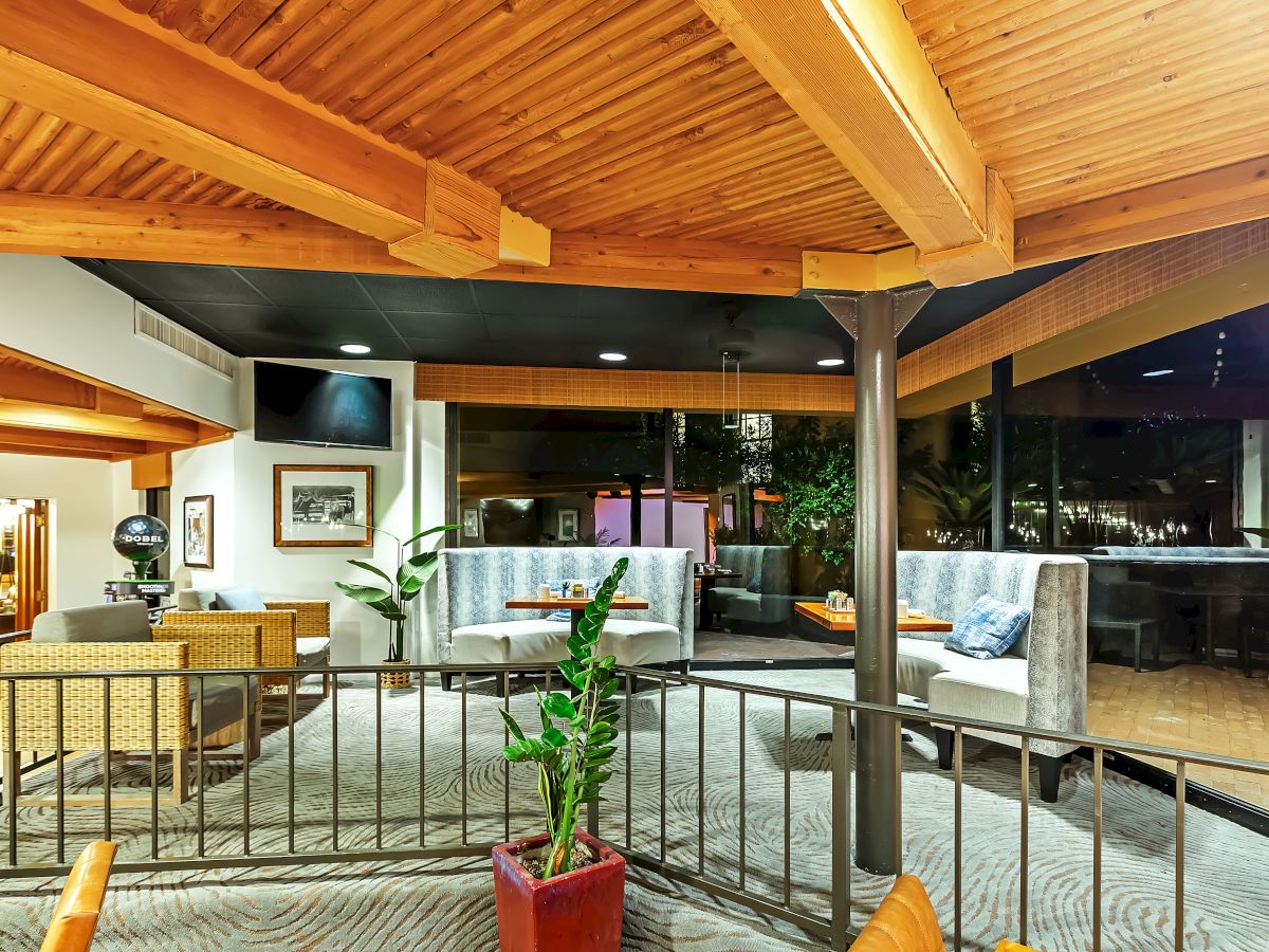 A modern interior space with wooden ceilings, a seating area, plants, and a TV on the wall, featuring large windows and cozy booths.