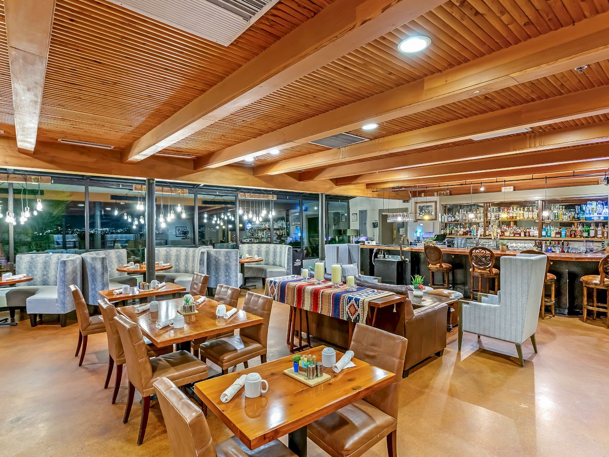 An inviting restaurant interior featuring wooden beams, tables, chairs, booths, and a well-stocked bar with a warm ambiance.