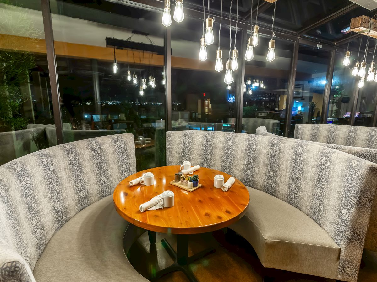 A round table with utensils and napkins is surrounded by a curved sofa in a well-lit restaurant with hanging lights and large windows.