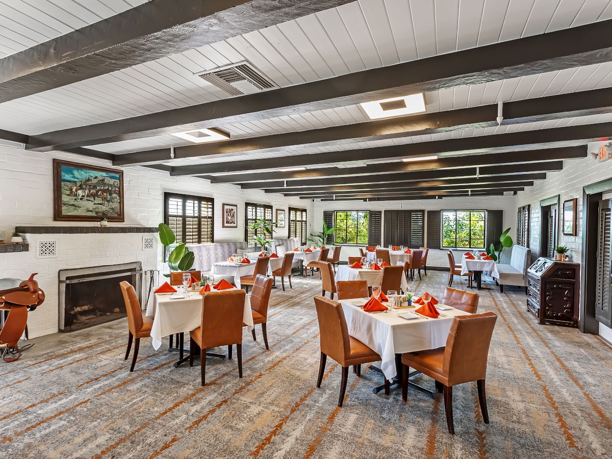 A spacious dining room with neatly arranged tables and chairs, set with white tablecloths and red napkins, featuring large windows and elegant decor.