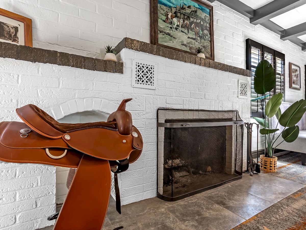 This image shows a living room with a brick fireplace, a horse saddle on a stand, plants, and artwork on the wall.