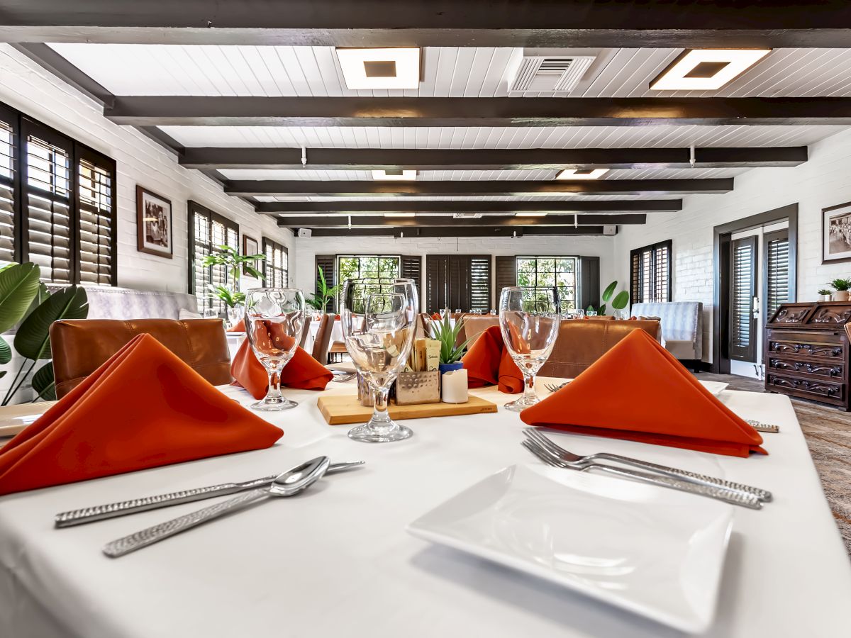 A restaurant dining room with white tablecloths, orange napkins, silverware, and wine glasses, with large windows and decorative plants.