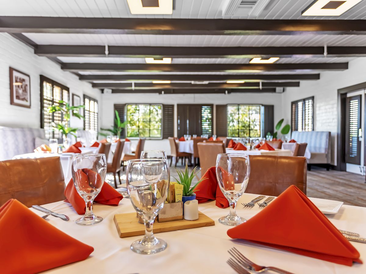 A well-lit restaurant with set tables, featuring orange napkins, glassware, and potted plants on each table, ready for guests.