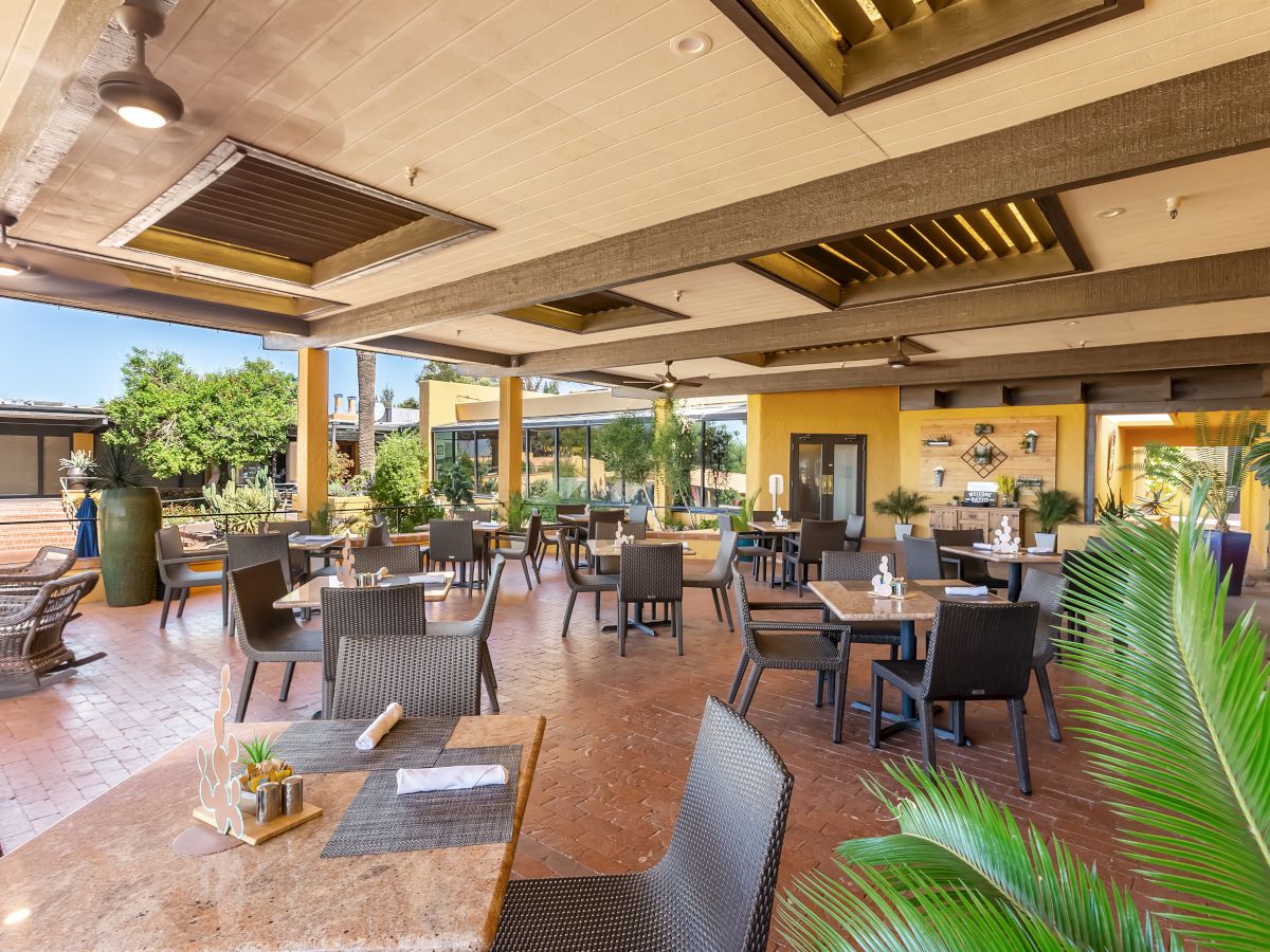 The image shows a spacious, partially outdoor dining area with several tables and chairs, surrounded by plants and well-lit with natural light.