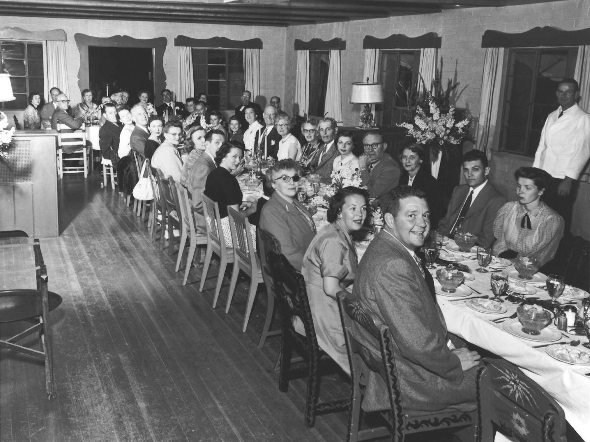 A large group of people seated at long tables in a dining room, dressed formally, possibly for a banquet or gathering.