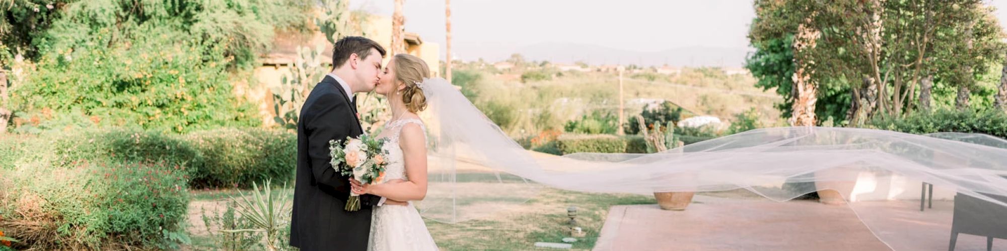 A bride and groom share a kiss outdoors, with her veil flowing in the wind. The background has trees and a clear sunny sky.