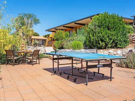 The image shows an outdoor patio area with a ping pong table and a dining table with chairs, surrounded by greenery and a building in the background.