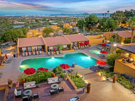 An aerial view of a resort featuring a central pool with lounge chairs, red umbrellas, outdoor seating, and surrounding buildings.