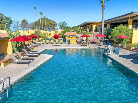 The image shows a luxurious outdoor swimming pool surrounded by lounge chairs and red umbrellas at a resort, with a modern building in the background.