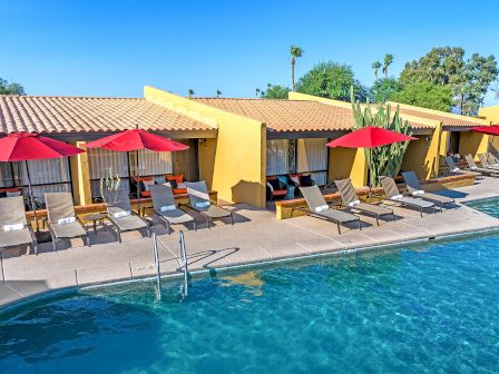 A row of poolside loungers with red umbrellas and modern villas in the background, set against a clear blue sky and surrounded by tropical plants.