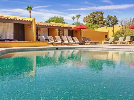 A serene poolside scene with lounge chairs, umbrellas, and a yellow building in the background, surrounded by palm trees and clear skies.