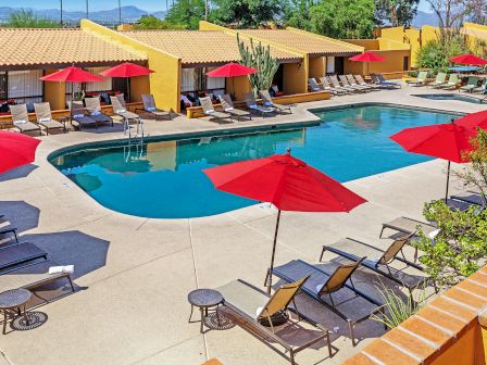 This image shows a tranquil outdoor pool area with lounge chairs, red umbrellas, and surrounding yellow buildings under a clear sky.