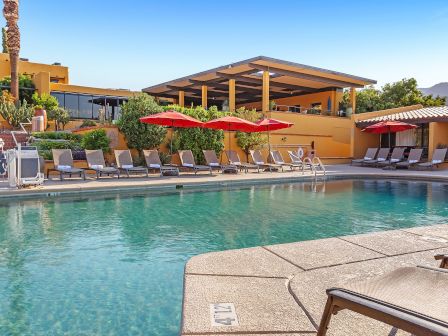 This image shows a serene outdoor pool area with lounge chairs, red umbrellas, and a large covered patio. The setting is sunny and inviting.