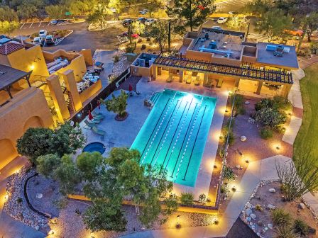 An aerial view of a brightly lit outdoor swimming pool surrounded by buildings and landscaped areas at dusk, among what appears to be a desert setting.