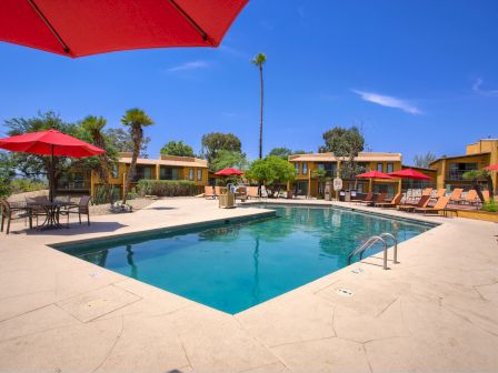 The image shows an outdoor pool area with red umbrellas, lounge chairs, and surrounding buildings under a clear blue sky.