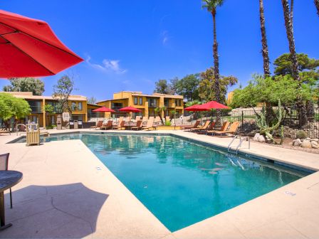 The image shows an outdoor pool area with lounge chairs, red umbrellas, and trees, surrounded by residential buildings and a clear blue sky.