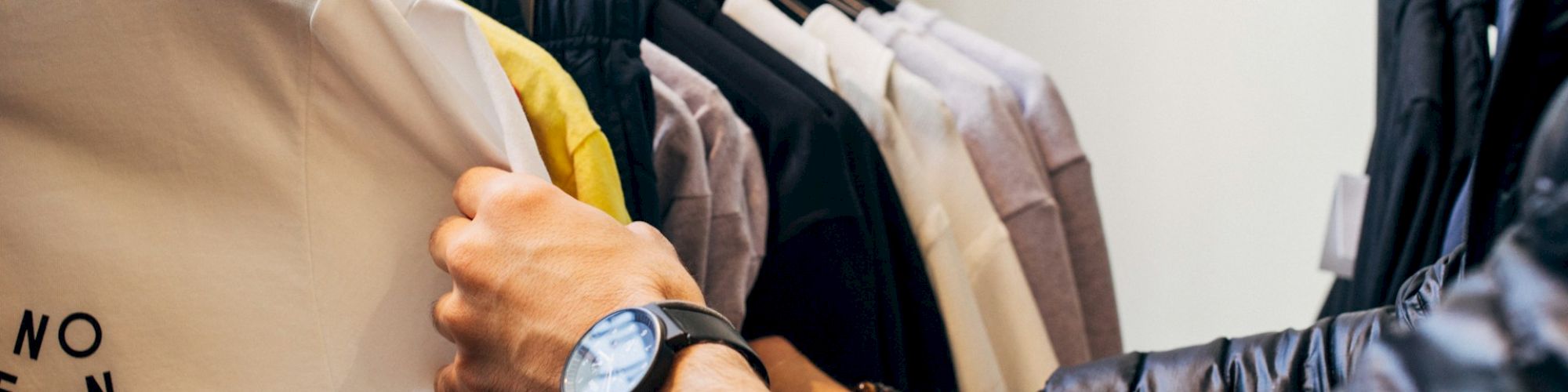 A person is browsing through clothing on a rack, holding a white T-shirt with "NO FUN" printed on it. The person has a watch on their wrist.