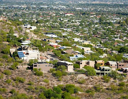 An arid landscape with a mix of desert vegetation, modern houses on a hillside, and a sprawling cityscape in the background, possibly in a desert region.