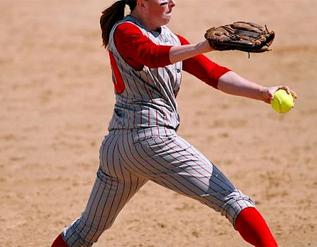 A softball player is in mid-pitch, holding a yellow softball in her right hand and wearing a gray and red uniform on a sandy field.