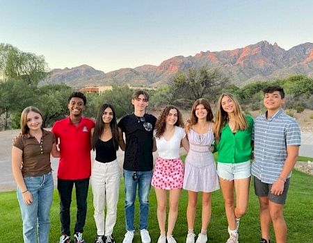 A group of eight young people stands together outdoors with a mountainous landscape in the background, smiling at the camera.