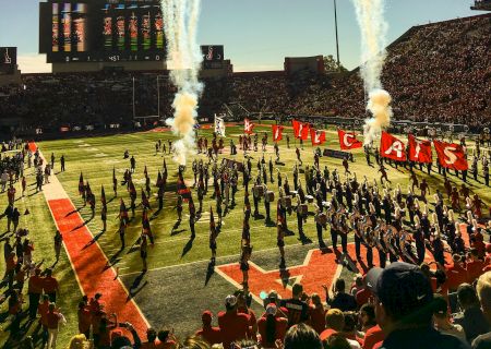 This image shows a football field with a marching band performing, fireworks in the sky, and a stadium filled with spectators during the daytime.