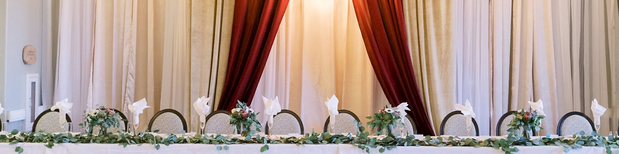 A long, elegantly decorated table set for an event, with chairs, white linens, folded napkins, greenery, and string lights above.