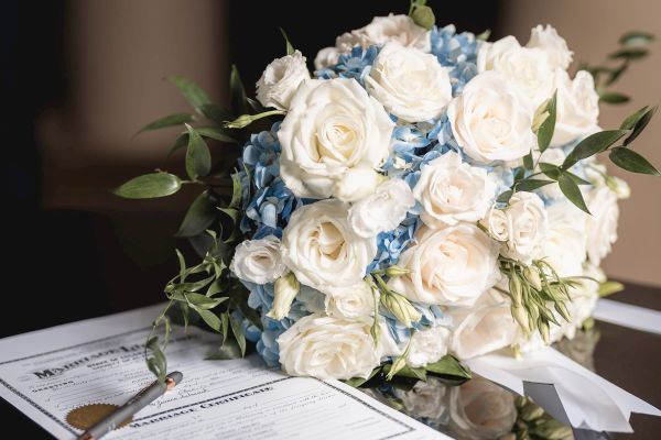 A bouquet of white and blue flowers is placed beside a document with a pen. The bouquet and document are reflected on a black surface.