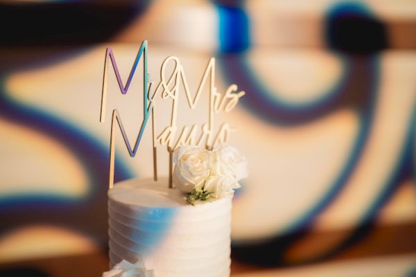 A white wedding cake with "Mr & Mrs" topper and white flowers against a blurred vibrant background.