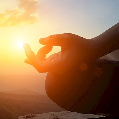 A person is meditating outdoors at sunset, with fingers forming a mudra gesture, against a scenic background with the sun setting behind them.