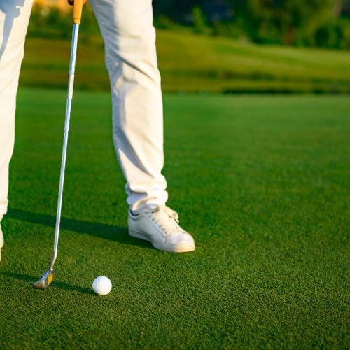 A person in white pants and shoes is on a golf course, preparing to putt a golf ball with a putter.