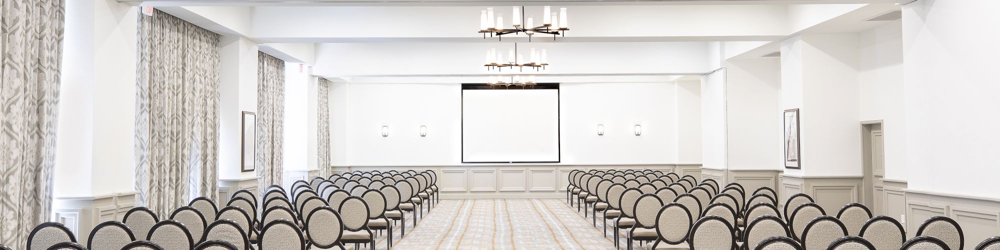 The image shows a modern conference room with rows of chairs facing a screen, chandeliers overhead, and light-colored decor and carpet.