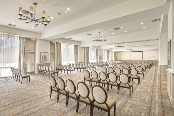 An empty conference room with rows of chairs facing a projector screen, large windows, chandeliers, and a neutral color scheme.