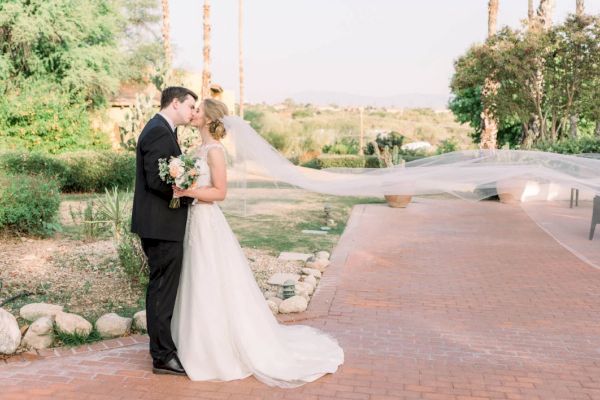 A bride and groom are kissing outdoors, with the bride's veil flowing in the breeze, surrounded by greenery and a brick path.