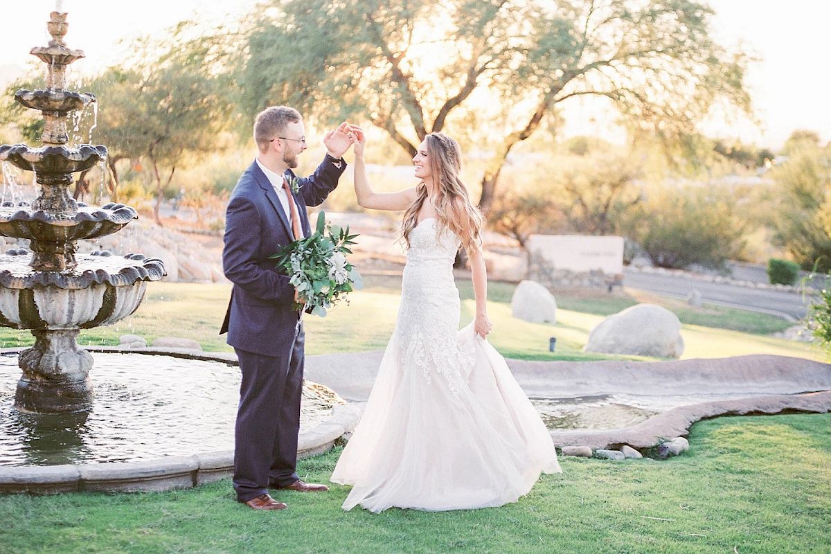 A couple, dressed formally, dances near a fountain in a park with trees and rocks in the background. The scene appears to be during sunset.