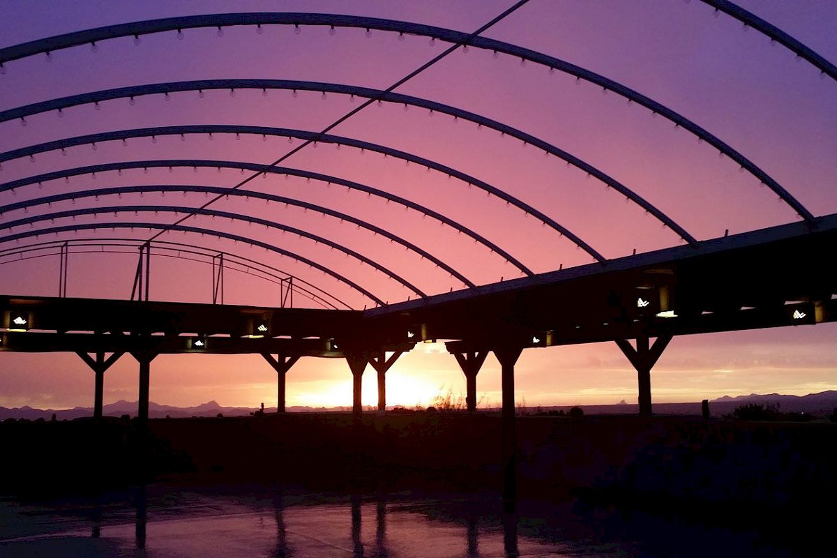 A structure with curved beams under a purple sunset sky, possibly an open-air venue or barn, silhouetted against the light, with mountains in the background.