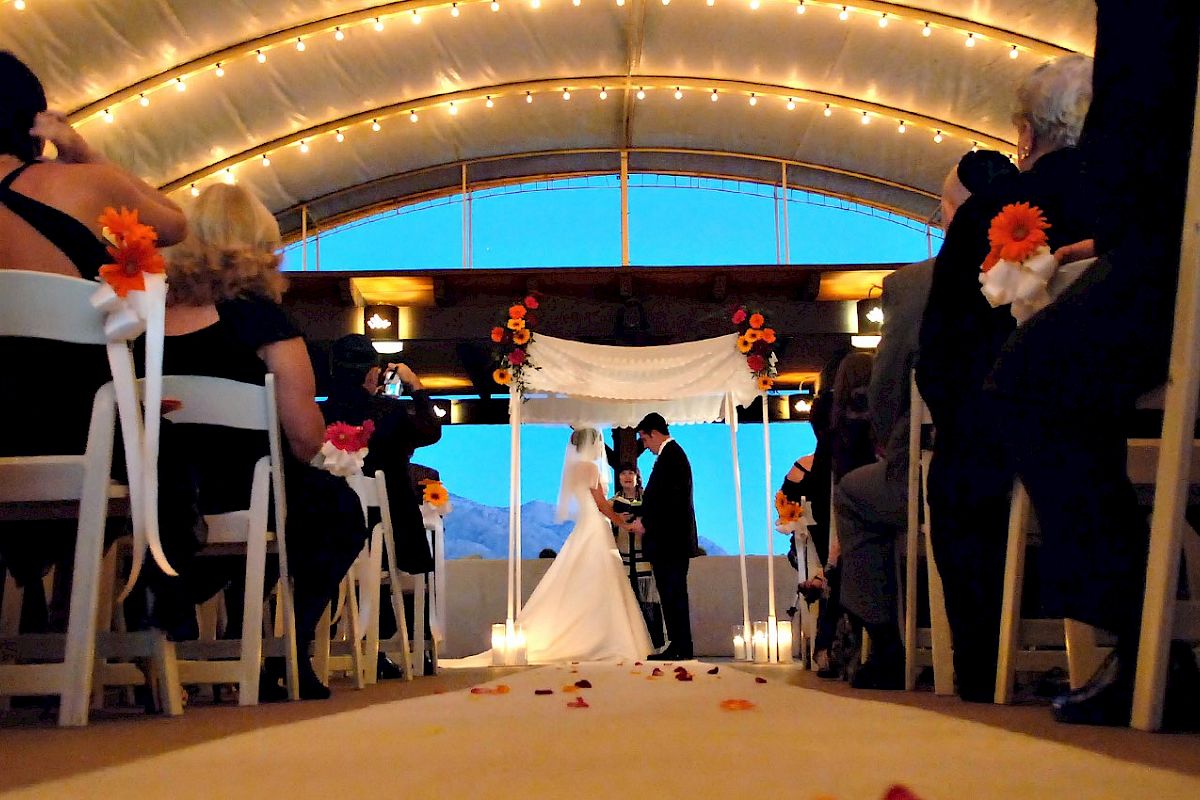This image shows an indoor wedding ceremony with a bride and groom under an archway, surrounded by guests seated on decorated chairs.