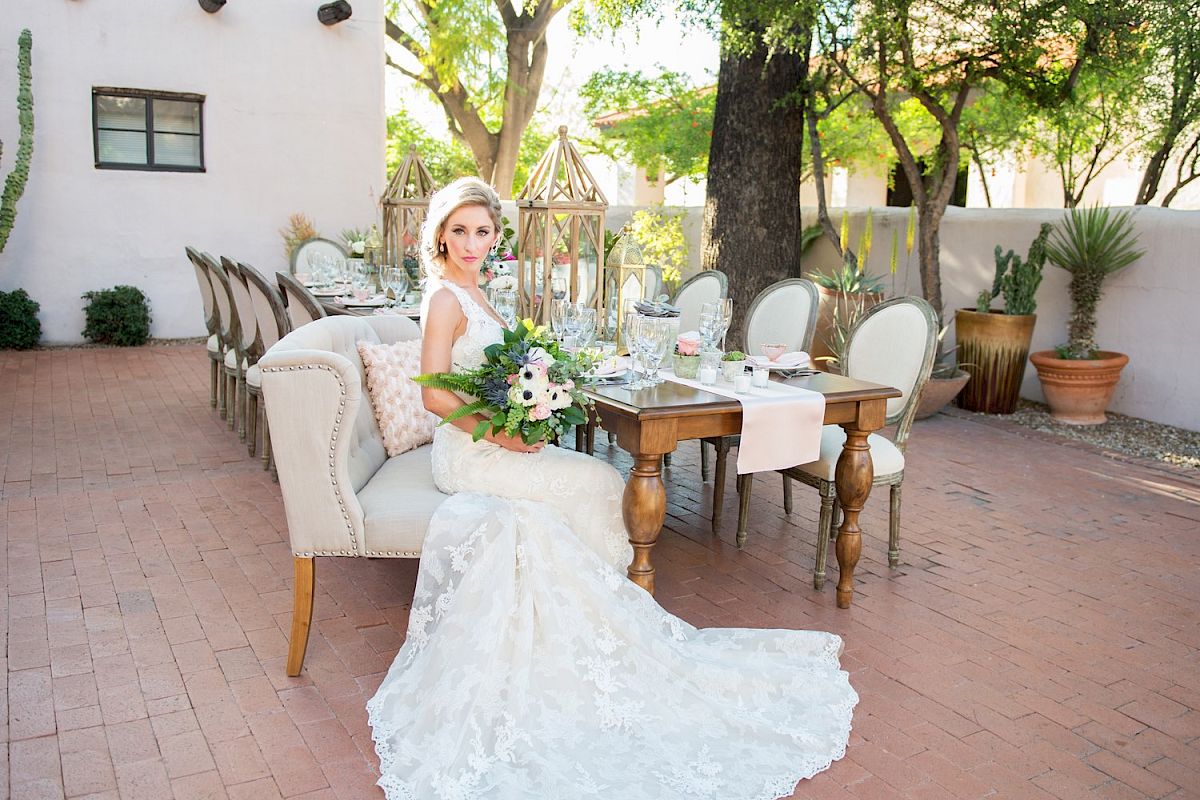A bride in a wedding dress sits on a sofa outdoors with a decorated dining table and chairs set up, surrounded by greenery and plants.
