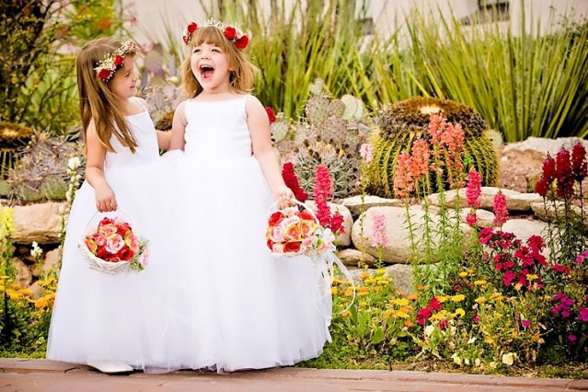 Two young girls in white dresses and flower crowns hold baskets of flowers in a colorful garden, with one girl laughing joyfully.