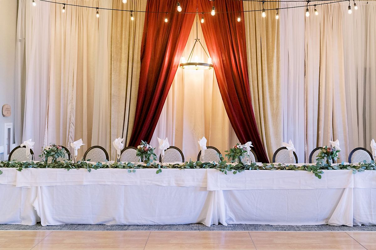 A long banquet table set with white linens, chairs, and floral arrangements is in front of draped curtains and string lights, creating an elegant ambiance.