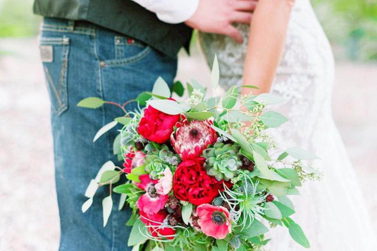 A couple dressed in wedding attire stands close, with the bride holding a bouquet of red flowers and greenery.