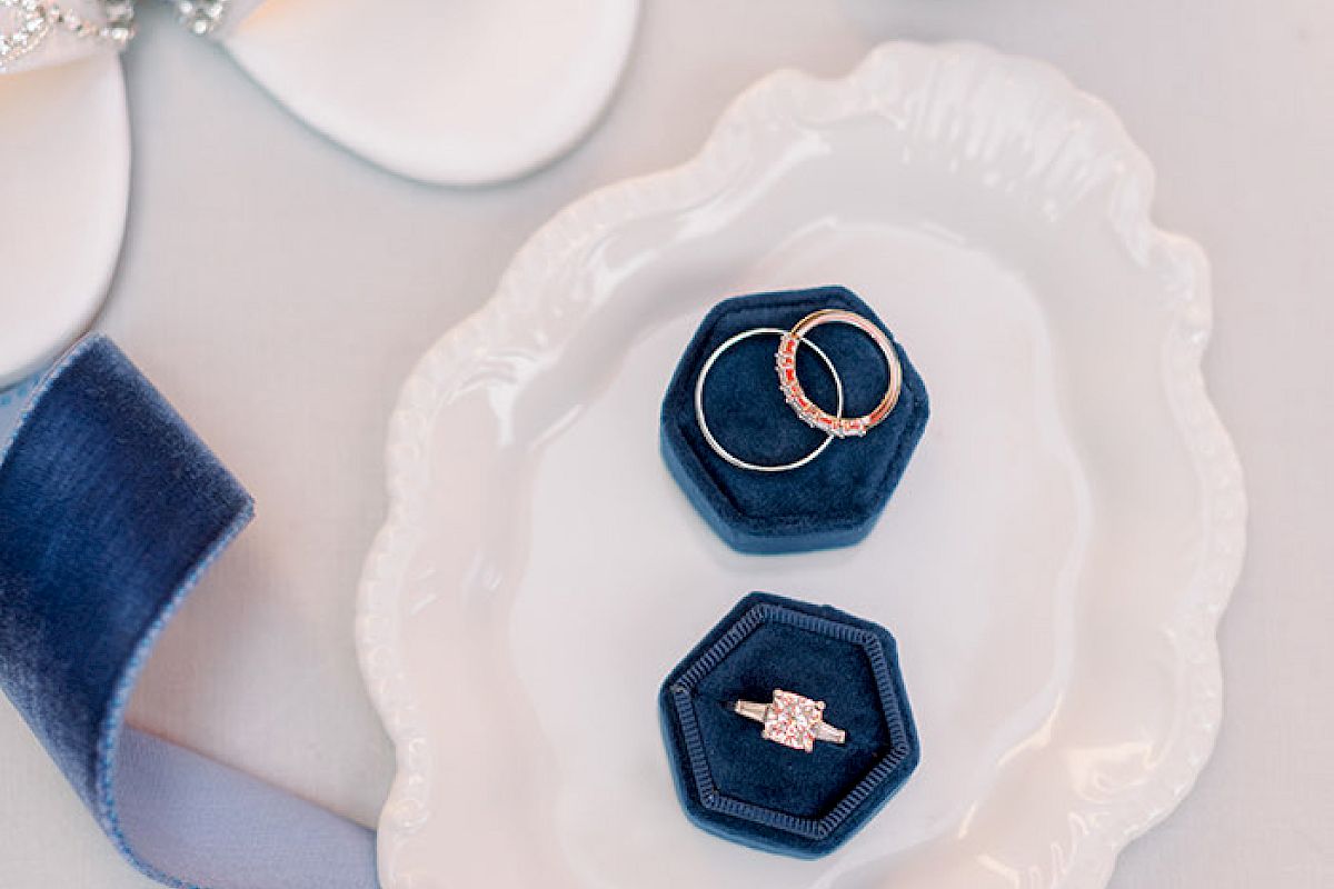 A decorative plate holds two ring boxes with rings, next to a shoe and a ribbon, and a round box holding a small jewelry piece in a gold container.