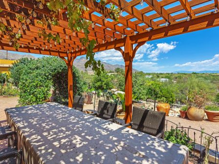 A wooden pergola with a covered table and chairs, overlooking a scenic view of greenery and mountains in the background under a clear blue sky.